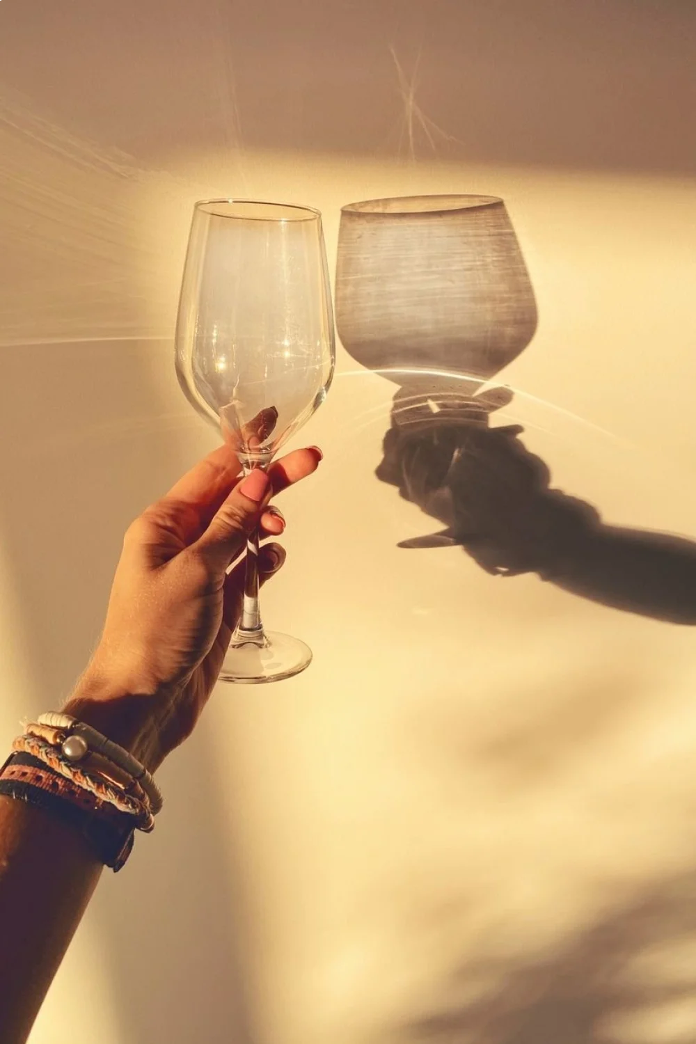 Hand holding a clear glass with a shadow on a light wall, featuring holographic pink polish on nails