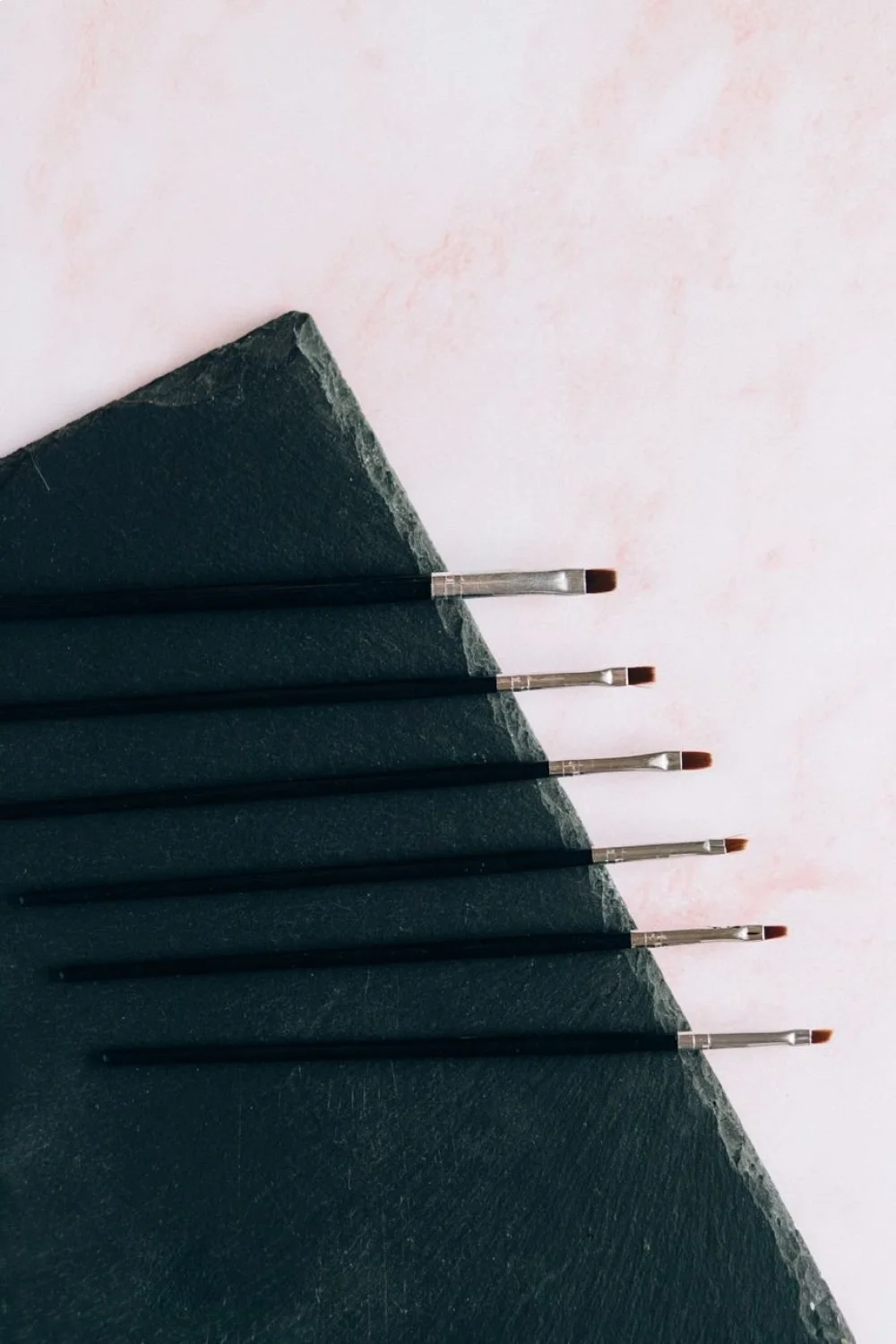 seven nail art brushes with angled tips on a dark slate background