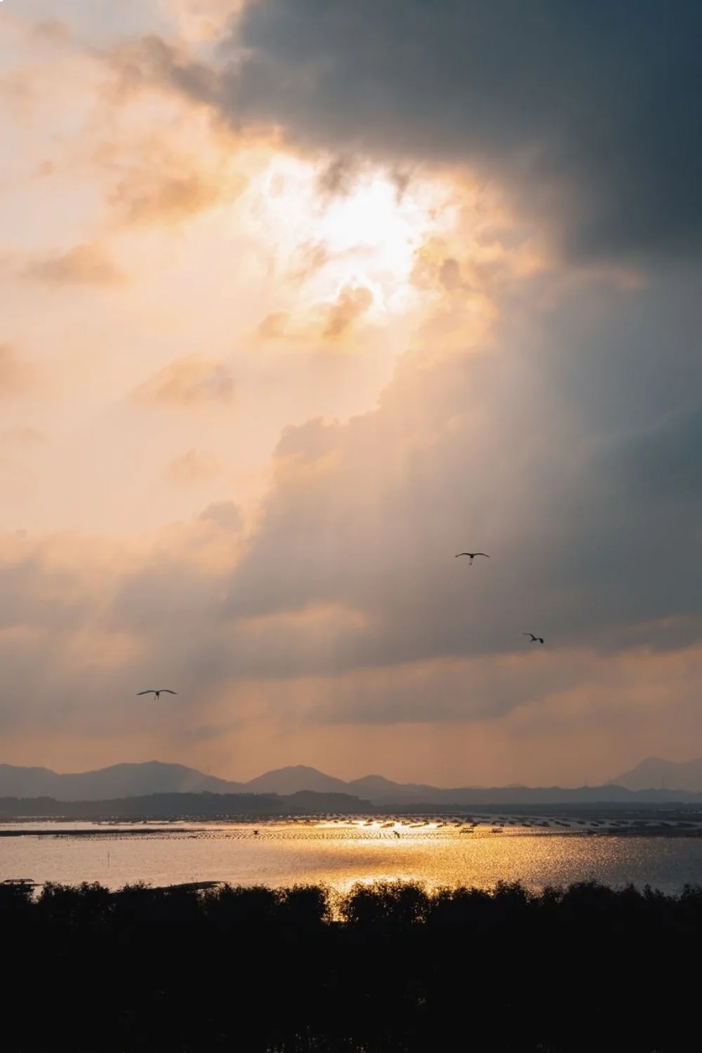 cloudy sky with rays of sunlight breaking through, reflecting on a calm water surface, mountains in the distance
